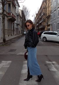 Veste en cuir noire, jupe longue en denim bleu clair avec une fente latérale, sac à main bordeaux et bottes à talons marron dans une rue.