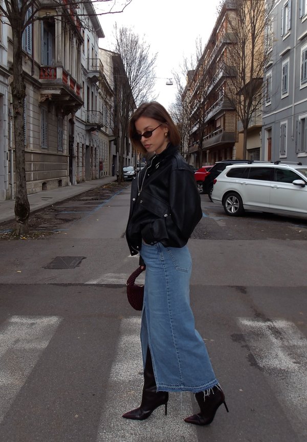 Chaqueta de cuero negro, falda larga de mezclilla azul claro con una abertura lateral, bolso burdeos y botas marrones de tacón alto en una calle.
