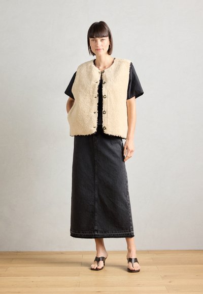 Chaleco de forro polar beige con cuello redondo y botones negros, llevado sobre una blusa negra de manga corta y combinado con una falda larga de mezclilla negra y sandalias.
