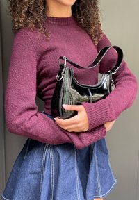 Femeie cu păr creț, purtând un pulover mov și o fustă albastră, ținând în mână o geantă mică, lucioasă, neagră, cu mâner superior, pe un fundal gri.