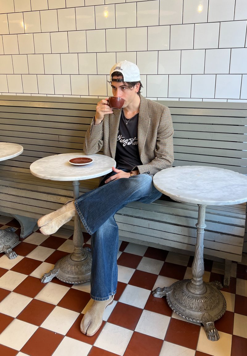 Homem com boné branco, blazer xadrez, jeans e botas bege a beber café num café com chão de azulejos e mesas redondas de mármore.