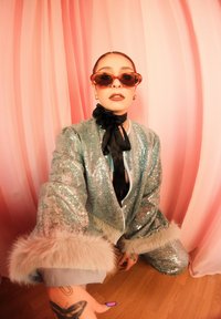 Costume vert à paillettes avec poignets en fausse fourrure, porté avec de grandes lunettes de soleil marron et un collier ruban noir. Fond rose, sol en bois.