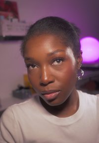 Jeune femme noire aux cheveux naturels tirés en arrière, portant des boucles d'oreilles argentées en forme de cœur et une chemise blanche, avec une lumière violette en arrière-plan.