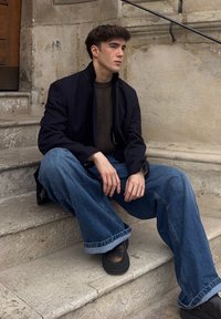 Modell iført en svart blazer over en brun genser, kombinert med løstsittende blå jeans og mørke sko, sittende på steintrapper.