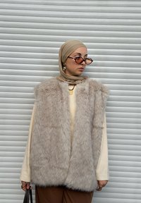 Gilet in pelliccia sintetica di colore beige chiaro, superficie strutturata, design con apertura frontale, indossato sopra una maglietta color crema e abbinato a occhiali da sole color bronzo.