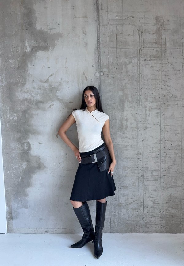 Kvinde iført hvid tætsiddende top, sort knælængde nederdel med stor bæltetaske og sorte knæhøje støvler, stående op ad tekstureret betonvæg.