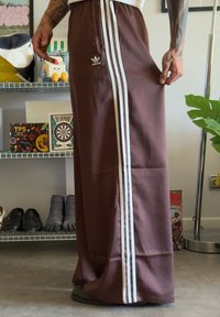 Marron tracksuit trousers with white side stripes, made of smooth fabric. Features an elastic waistband and an Adidas logo on the front.