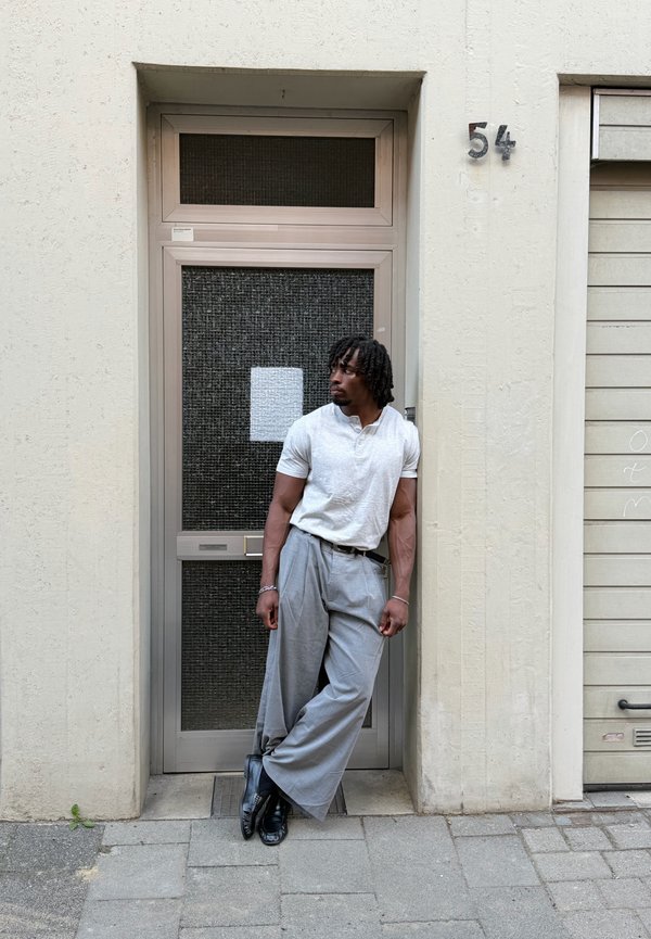 Homme portant une chemise blanche et un pantalon large gris s'appuie contre une porte numérotée 54 sur un trottoir pavé en milieu urbain.