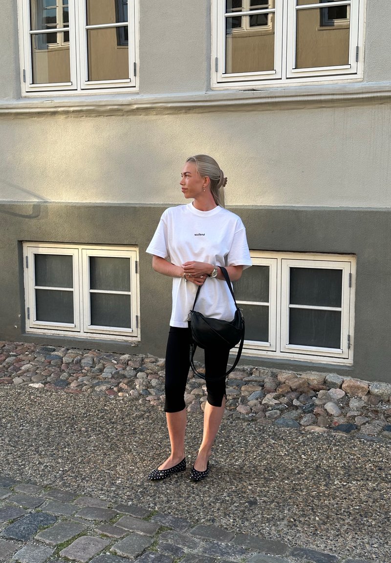 Λευκό oversized t-shirt με κείμενο "Holland", μαύρα cropped κολάν και μαύρα παπούτσια με πουά. Μαύρη τσάντα ώμου, απαλό μαλλί, αστική σκηνή.