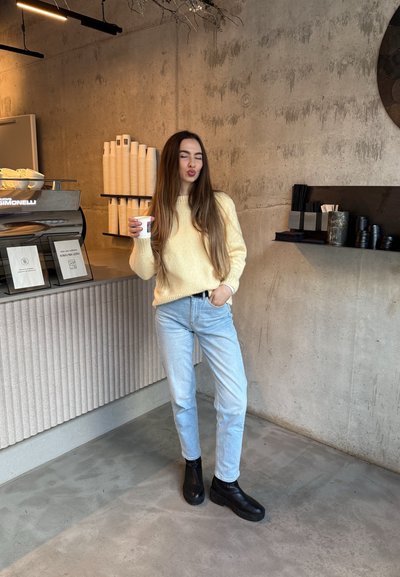 Joven mujer con suéter amarillo y jeans sostiene una taza de café, de pie en un café minimalista con paredes de concreto y tazas apiladas.