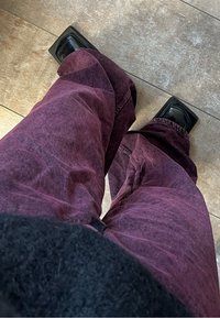 Svarta plattformsskor med fyrkantig tå som bärs med lösa, strukturerade, mörklila jeans. Golvet är ljust och plattlagt.