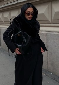 Fekete kabát szőrme sallanggal, sötét kapucnis pulóverrel és barna tónusú napszem�üveggel. Elegáns fekete táskát tart hosszú pántokkal. Városi környezetben.