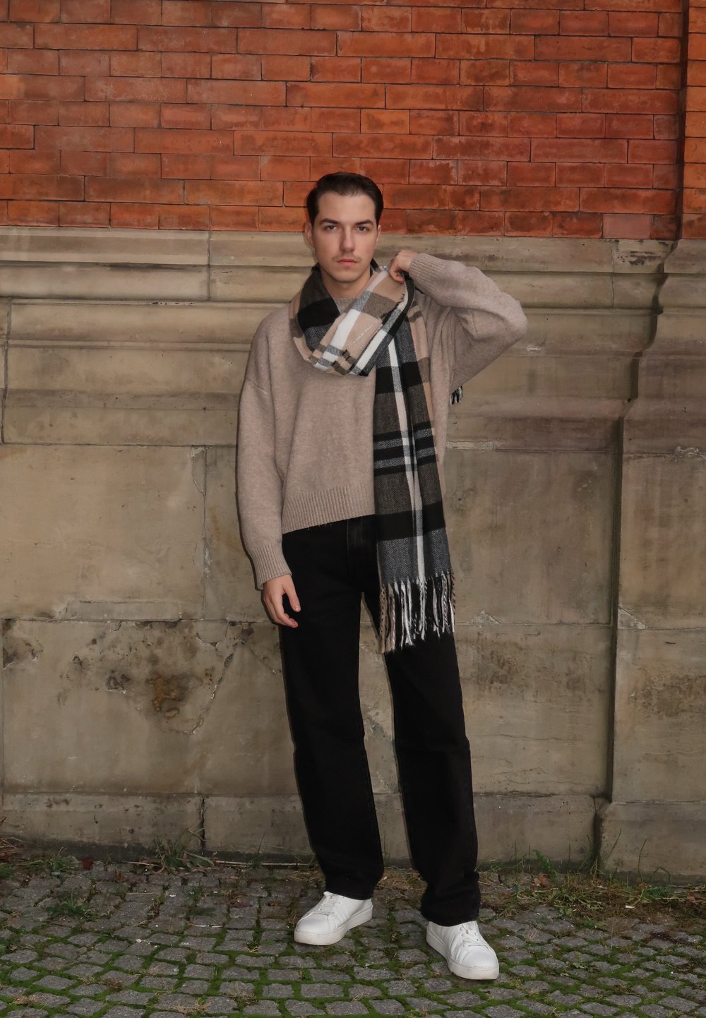 Pull en tricot beige, écharpe à carreaux gris et noirs, pantalon noir et baskets blanches. Debout contre un mur en briques.