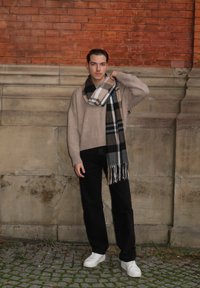 Pull en tricot beige, écharpe à carreaux gris et noirs, pantalon noir et baskets blanches. Debout contre un mur en briques.