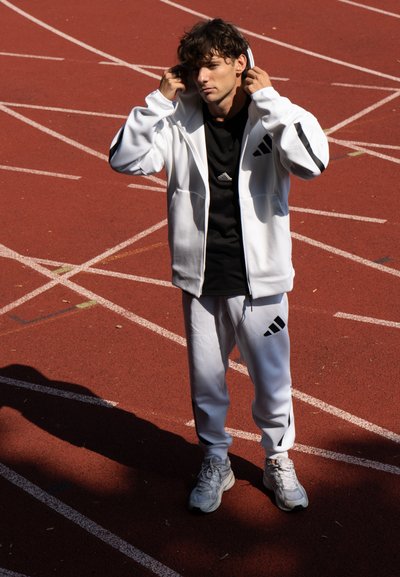 Chaqueta y pantalones deportivos blancos con acentos negros y tres rayas; camiseta negra debajo; de pie sobre una pista roja.