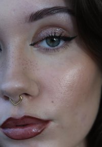 Close-up of a face featuring light freckling, a gold heart-shaped nose ring, glistening eyeshadow, and deep red glossy lips.