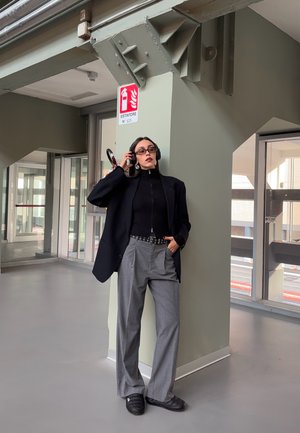 Mujer con pantalones grises y blazer negro se apoya en un pilar en interiores, lleva gafas y auriculares, con una mano en el bolsillo y la otra ajustando los auriculares.