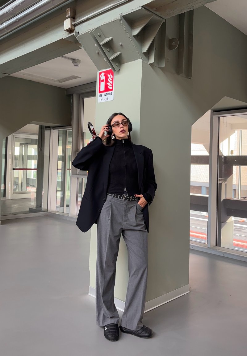 Frau in grauen Hosen und schwarzem Blazer lehnt drinnen an einer Säule, trägt eine Brille und Kopfhörer, hat eine Hand in der Tasche und die andere passt die Kopfhörer an.