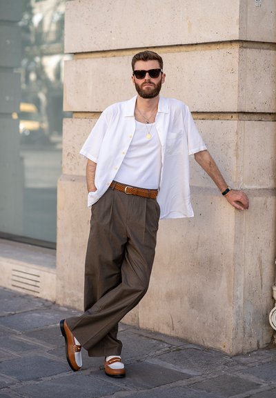 Camisa blanca de manga corta sobre una camiseta blanca, pantalones sueltos marrones y zapatos marrones y blancos. Los accesorios incluyen gafas de sol y un collar.