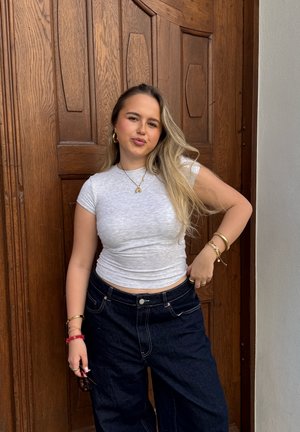 Graues, enges T-Shirt mit kurzen Ärmeln, hochtaillierte, dunkle Jeans im weit geschnittenen Stil. Goldene Creolen und geschichtete Armbänder, die an einer Holztür stehen.