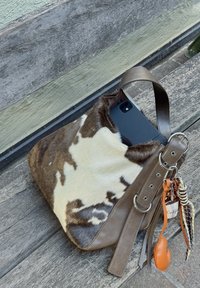 Bolso de hombro de piel de vaca marrón y blanca con correa de cuero y charms en el llavero, que contiene un smartphone negro, descansando sobre un banco de madera.