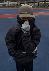 Fekete vízálló kapucnis dzseki, szürke kesztyűk és világos sapka; sportkörnyezetben viselve, a háttérben kék és piros pálya.