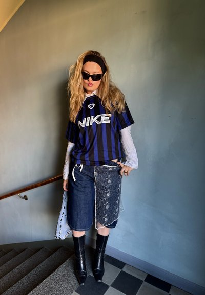 Jersey de Nike a rayas negras y azules, superpuesto sobre una camiseta de manga larga blanca. Pantalones cortos de mezclilla con una capa transparente, combinados con botas negras de tacón alto.