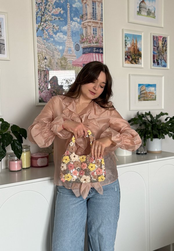 Femme en blouse rose transparente et jeans tenant un petit sac à main floral à sequins, debout devant un cabinet blanc et des affiches de voyage encadrées.