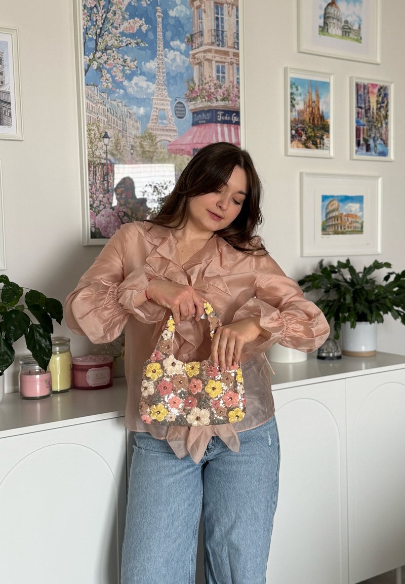 Femme en blouse rose transparente et jeans, tenant un petit sac à main floral à sequins, debout devant un meuble blanc et des affiches de voyage encadrées.
