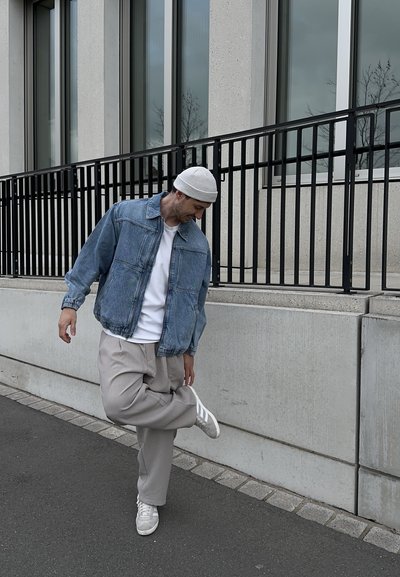 Hombre con chaqueta de mezclilla y gorro atándose el zapato mientras está de pie en una acera junto a un edificio con grandes ventanales y una barandilla de metal.