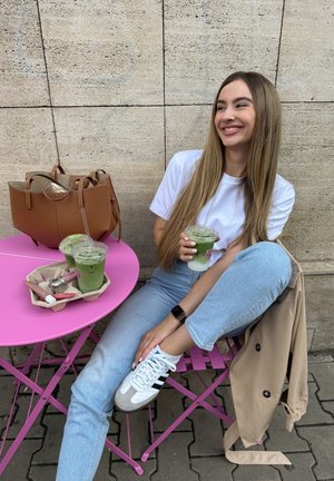Llevando una camiseta blanca y jeans azules, sosteniendo una bebida verde. A su lado hay una bolsa marrón y una mesa rosa con dos bebidas y cosméticos.