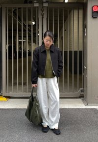 Svart bomberjacka, olivgrön tröja, glansiga vita vida byxor, svart skor och en olivfärgad väska, mot en bakgrund med grind.