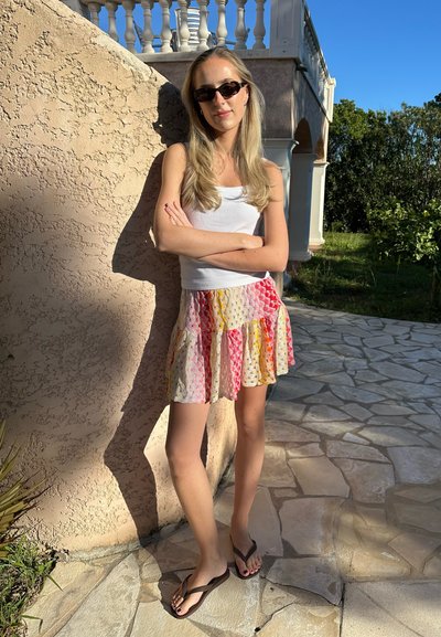 Camiseta blanca sin mangas, falda acampanada colorida con patrones en rosa, amarillo y naranja, y chanclas marrones. Escenario al aire libre con luz natural.