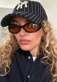 Kvinnan med lockigt hår bär en randig keps från New York Yankees, stora bärnstensfärgade solglasögon och en mörk jacka med knappar.