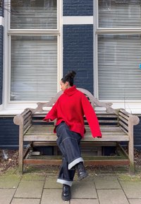 Maglione a collo alto a coste rosso, vestibilità oversize, indossato con jeans in denim scuri a gamba larga e stivali neri. Sfondo con panchina di legno e muro di mattoni blu.