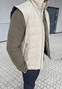 Persoană purtând o vestă matlasată bej peste un pulover tricotat verde măsliniu, cu pantaloni negri, stând pe un paviment din gresie lângă o clădire gri.