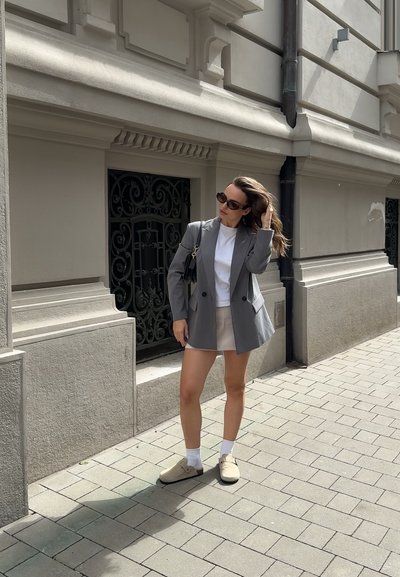 Blazer gris sobre una camiseta blanca y una falda beige clara, combinado con zapatillas beige y calcetines blancos, frente a una pared de piedra con barandilla decorativa.