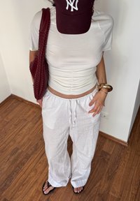Persona que lleva un top corto arrugado blanco y pantalones holgados blancos, chanclas negras, una gorra marrón con un logo blanco y lleva una bolsa de mimbre marrón.