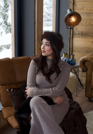 Mujer con atuendo gris y sombrero de piel sentada en un sofá marrón, mirando por una ventana nevada en una acogedora habitación de madera con iluminación cálida.