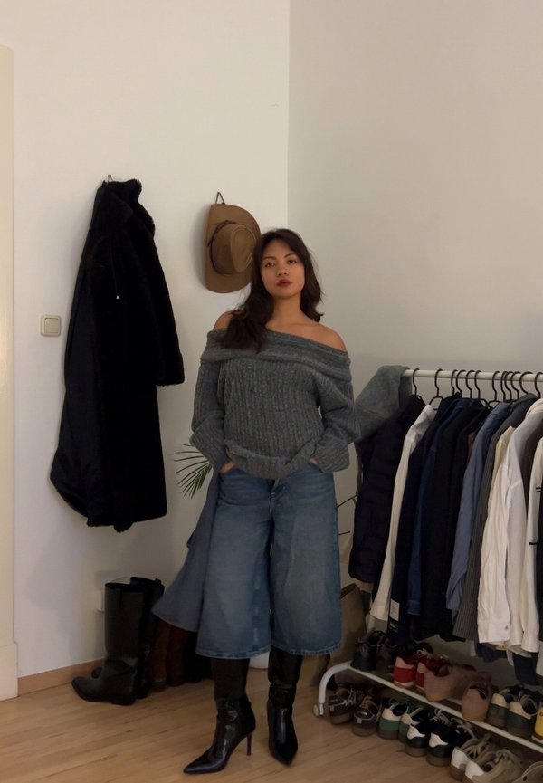 Nainen, joka pukeutuneena olkapäät paljastavaan harmaaseen villapaitaan, leveisiin farkkuihin ja mustiin kärjellisiin saappaisiin, seisoo huoneessa, jossa on vaatteita ja kenkiä naulakoissa.