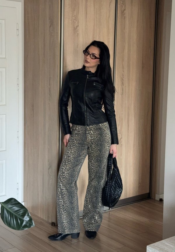 Donna con capelli lunghi e scuri indossa una giacca di pelle nera, pantaloni con stampa leopardata, scarpe nere con tacco, occhiali e tiene una borsa nera intrecciata al chiuso.