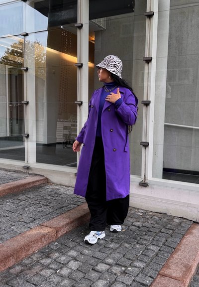 Persona vestida con un abrigo largo morado, pantalones negros, zapatillas blancas y un sombrero tipo bucket con rayas de cebra, de pie en una rampa de adoquines junto a ventanas de vidrio.