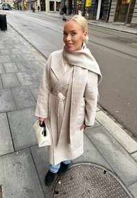 Bežni plašč z širokimi reverji in pasom, v kombinaciji s svetlo modrimi kavbojkami in črnimi gležnjarji. Drži malo belo torbico.
