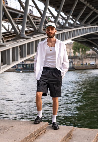 Camisa blanca de botones sobre una camiseta sin mangas blanca, pantalones cortos negros, calcetines blancos y zapatos negros. De pie junto al agua, bajo un puente.