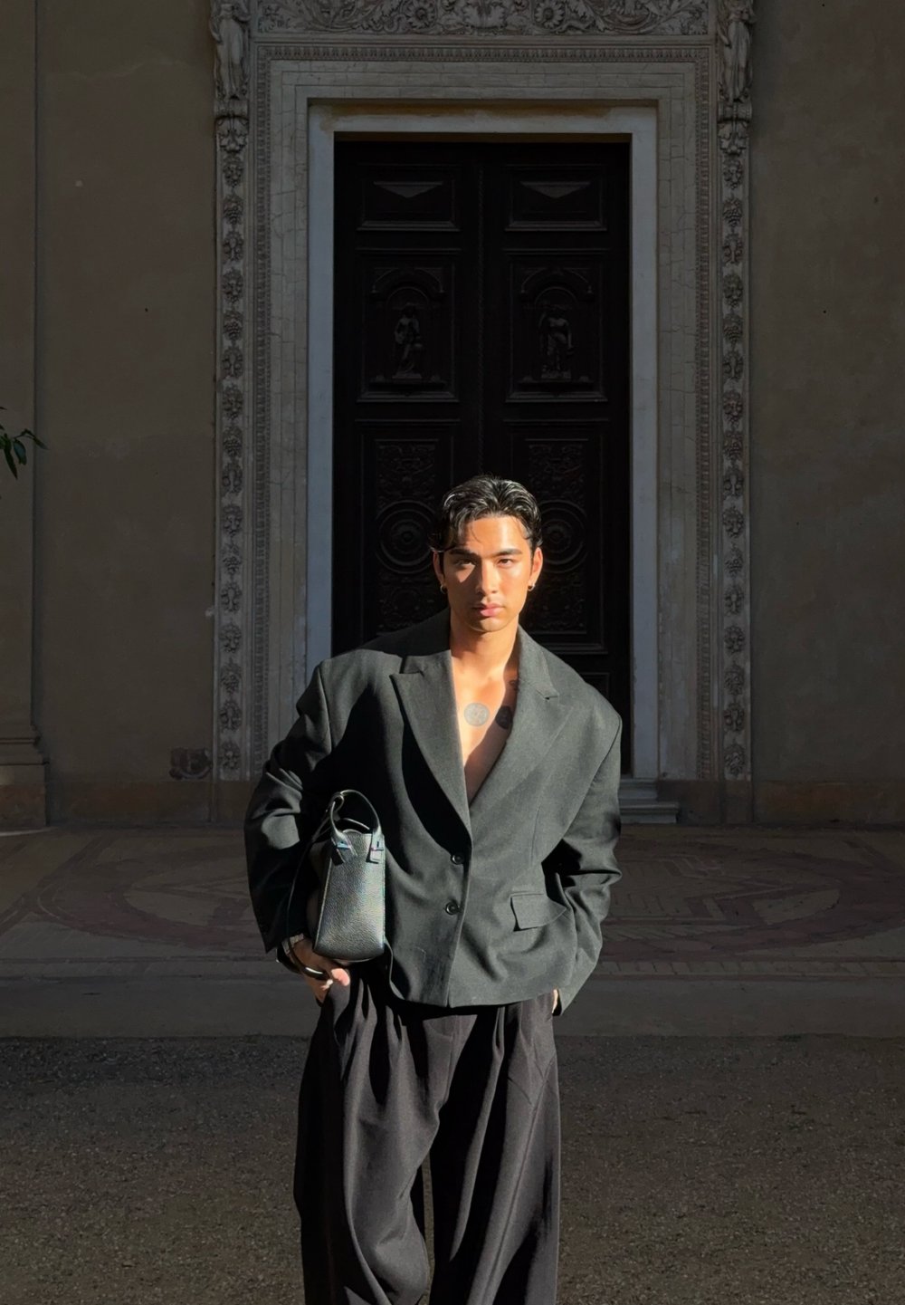 Junger Mann mit kurzen dunklen Haaren und Tattoos trägt einen übergroßen dunklen Blazer und weite Hosen, hält eine schwarze Tasche und steht vor einer kunstvoll verzierten Tür.