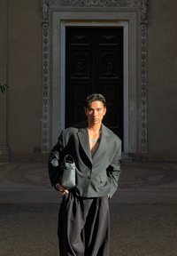 Junger Mann mit kurzen dunklen Haaren und Tattoos trägt einen übergroßen dunklen Blazer und weite Hosen, hält eine schwarze Tasche und steht vor einer kunstvoll verzierten Tür.