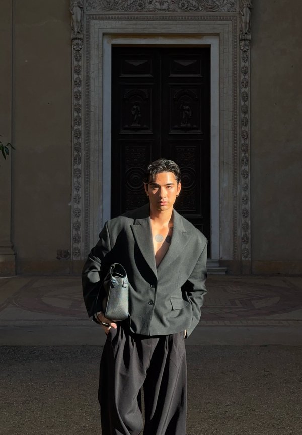 Joven con cabello corto y oscuro y tatuajes, viste un blazer oscuro de gran tamaño y pantalones anchos, sostiene un bolso negro, de pie frente a una puerta ornamentada.