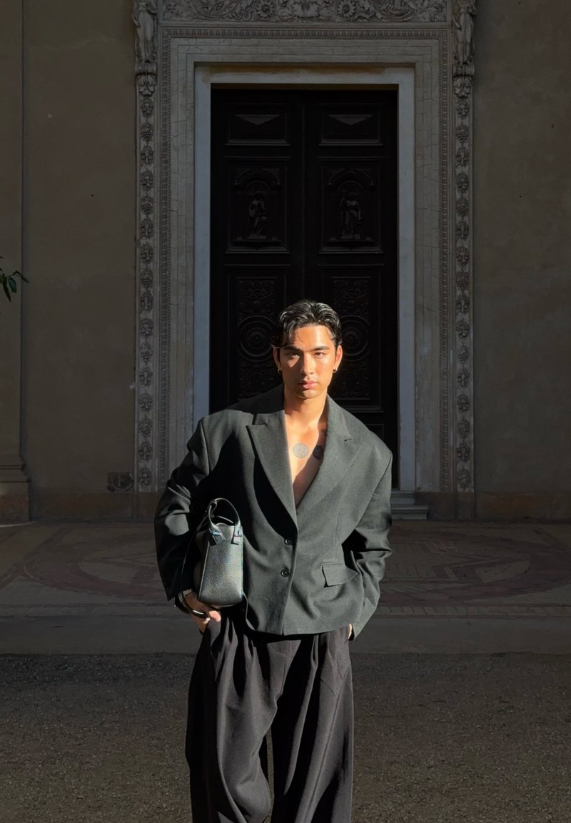 Junger Mann mit kurzem dunklem Haar und Tätowierungen trägt einen oversized dunklen Blazer und weite Hosen, hält eine schwarze Tasche und steht vor einer reich verzierten Tür.