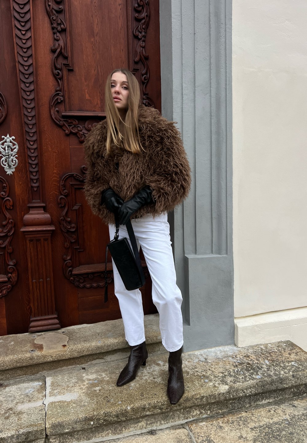 Femme aux longs cheveux blonds portant une veste en peluche marron, des gants noirs, un pantalon blanc et des bottines foncées, debout près d'une porte en bois sculpté.