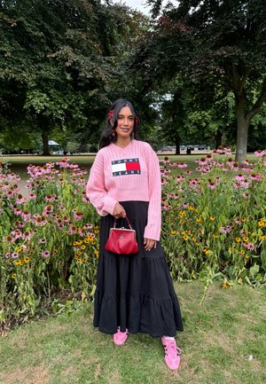 Suéter de punto rosa claro con el logo "TOMMY JEANS", falda larga negra con volantes, bolso rojo, zapatillas rosas, fondo floral con margaritas.